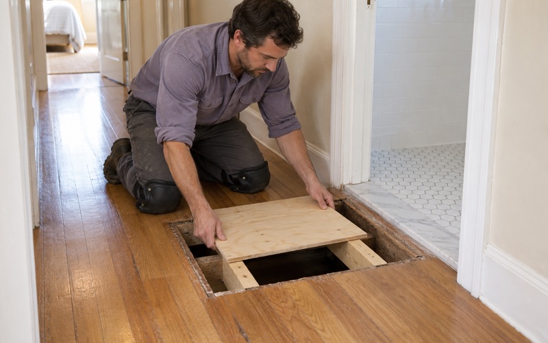 Worker lowering a new plywood subfloor patch onto the joists under an opened bathroom-doorway floor in a Lakewood house.